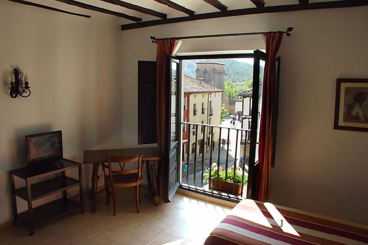 A room with a bed, a desk, a chair, and a balcony with a view of buildings and a mountain.