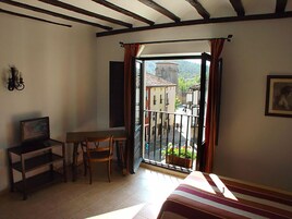 A room with a bed, a desk, a chair, and a balcony with a view of buildings and a mountain.