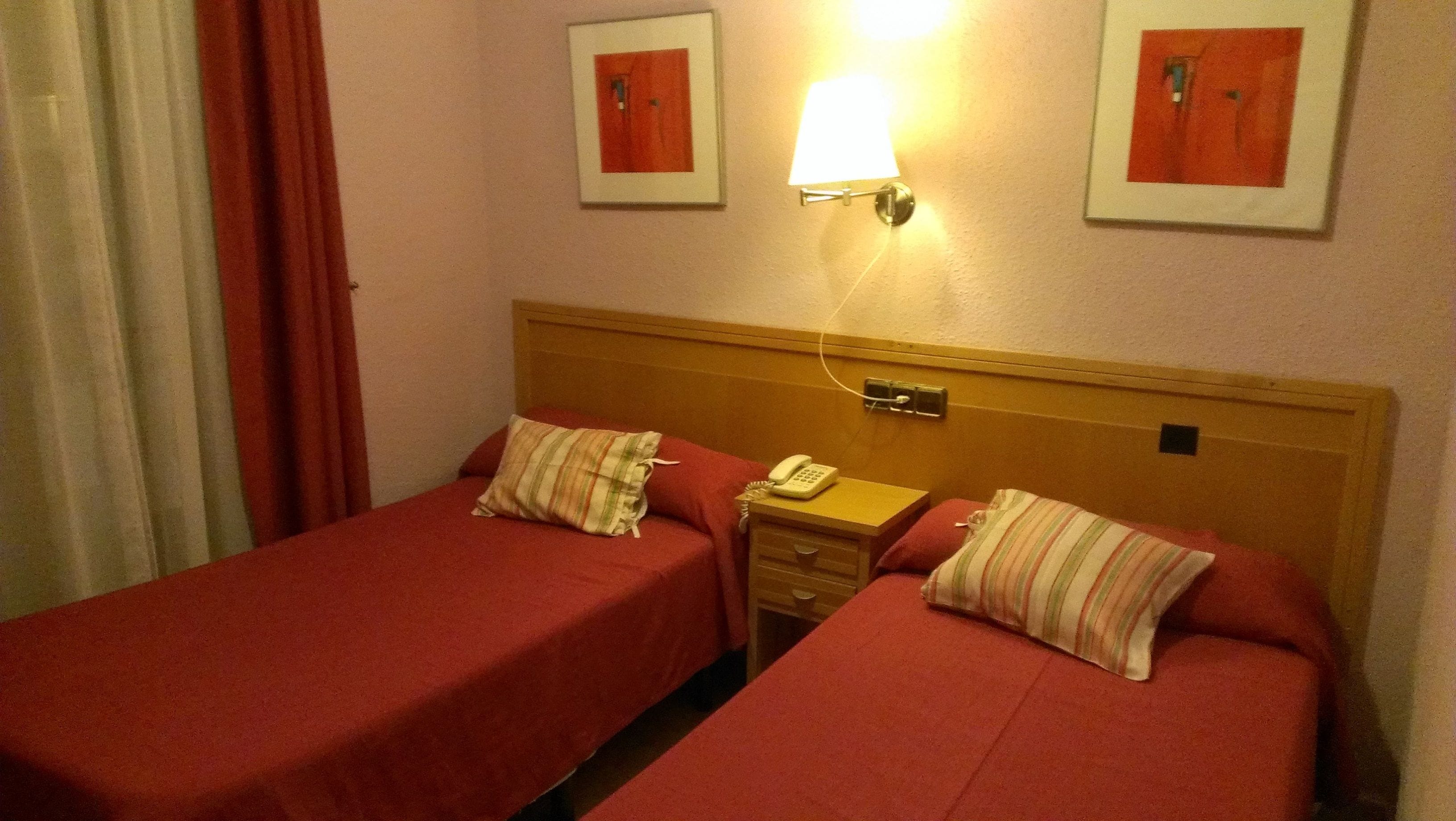 A hotel room with two single beds, each with a red bedspread and striped pillows. There is a bedside table with a lamp and a telephone. The room has a wooden headboard and a wall-mounted light fixture.