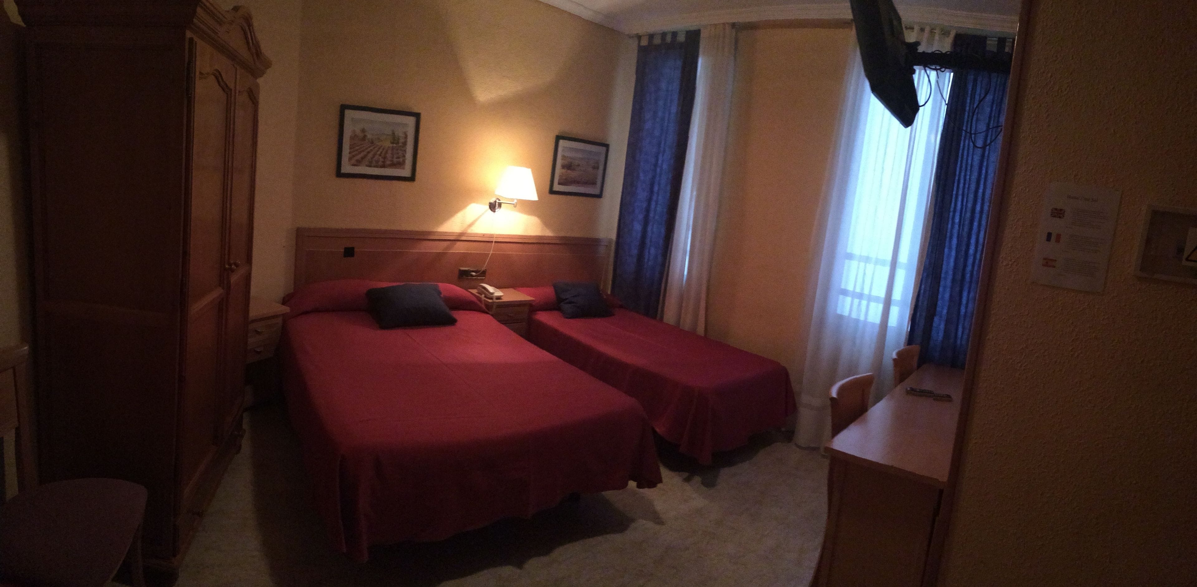 Une chambre d’hôtel avec deux lits simples, un bureau, une chaise, une lampe et une fenêtre avec des rideaux.