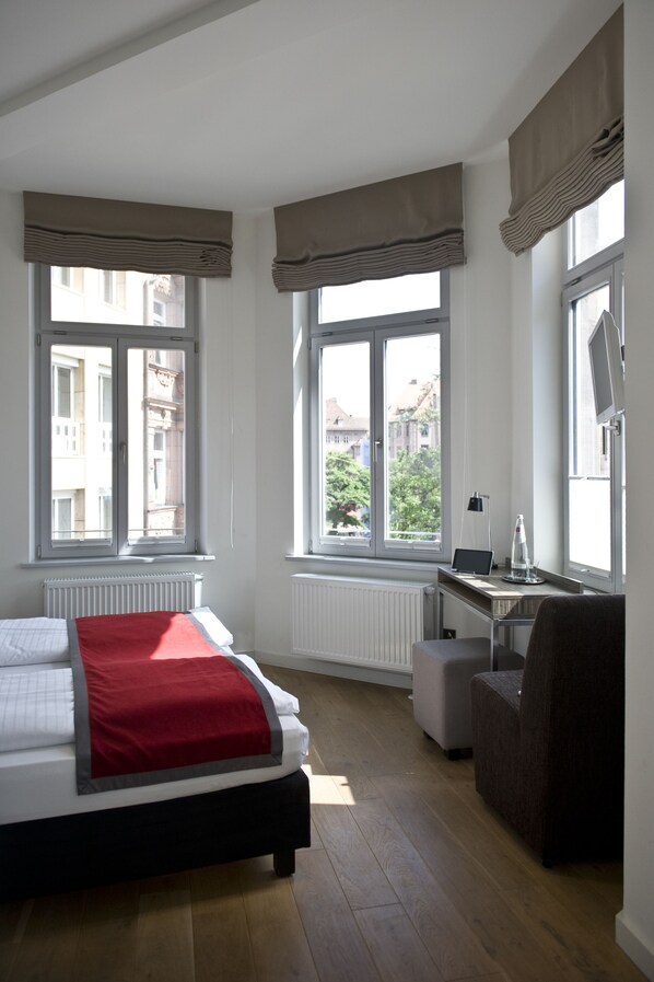 A bedroom with a bed, a desk, and a chair. There are windows with curtains, a radiator, and a view of buildings outside. - Gideon Designhotel (Nuremberg)
