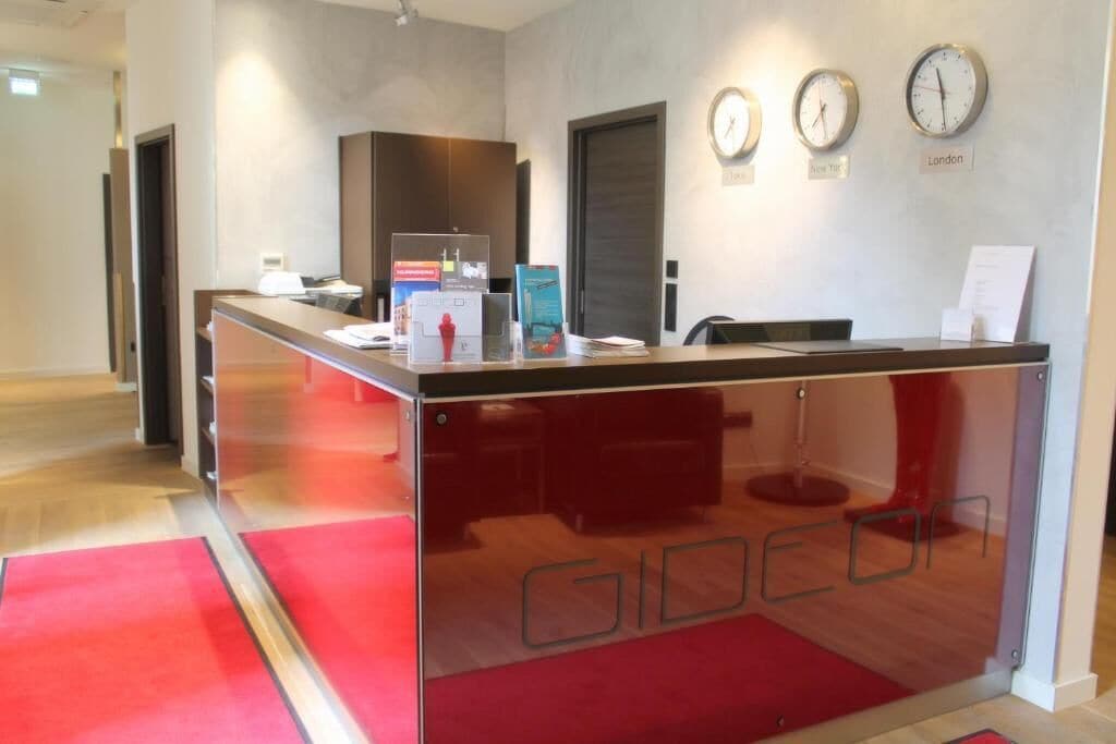 Un mostrador de recepción con un separador de vidrio, relojes que marcan diferentes husos horarios y una alfombra roja que conduce a la entrada.