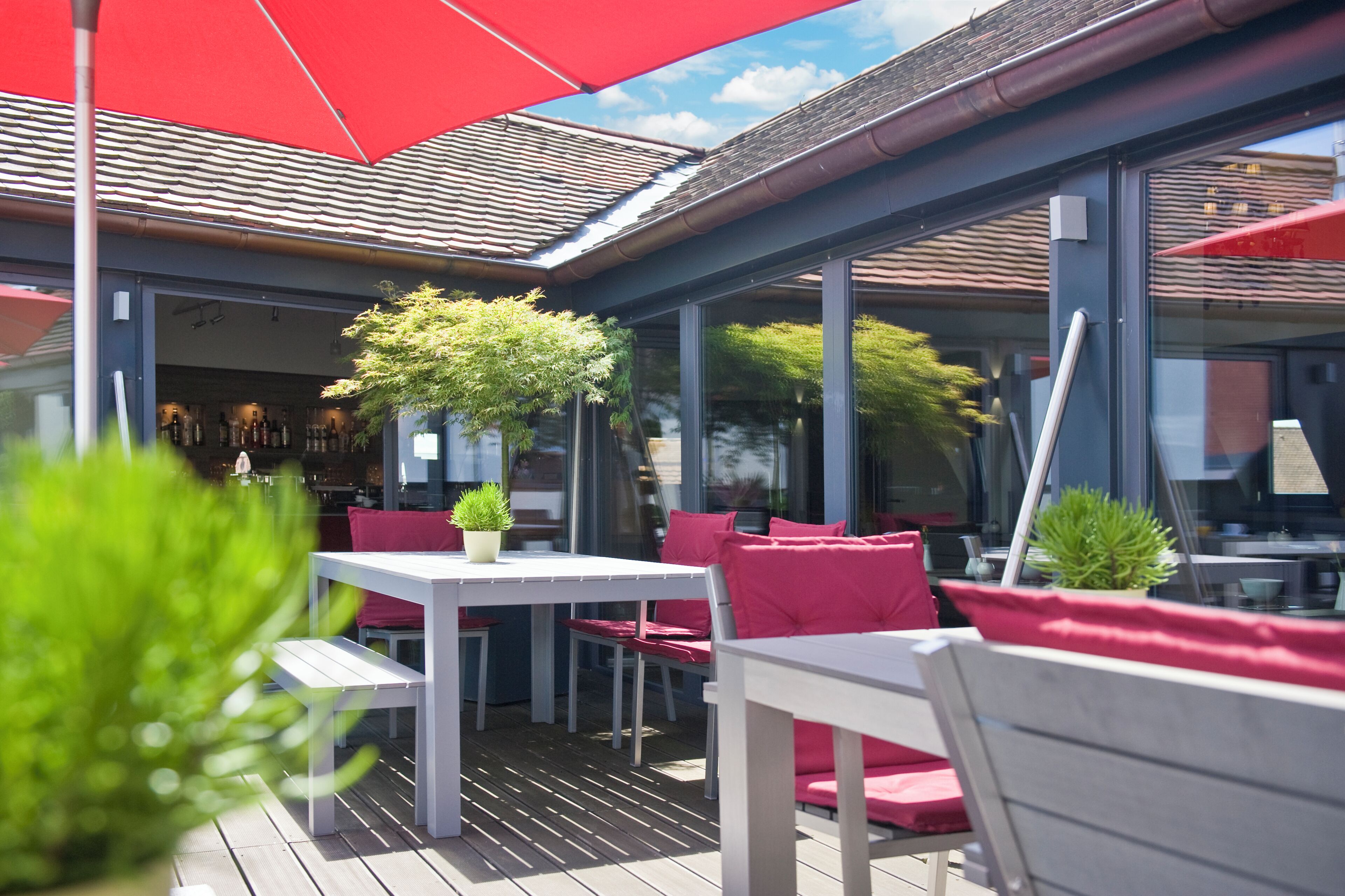 An outdoor patio with white tables and red chairs, a red umbrella, and potted plants.