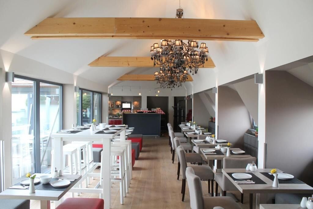 A modern restaurant with high ceilings, exposed wooden beams, and a central chandelier. Tables are neatly arranged with white chairs, and the space is well-lit with natural light from large windows.