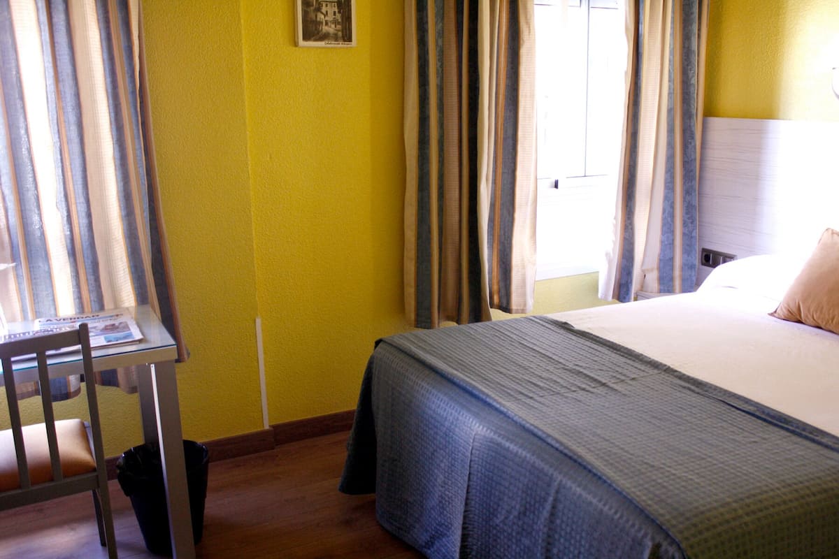 A hotel room with a bed, a chair, a table with a newspaper, and curtains.