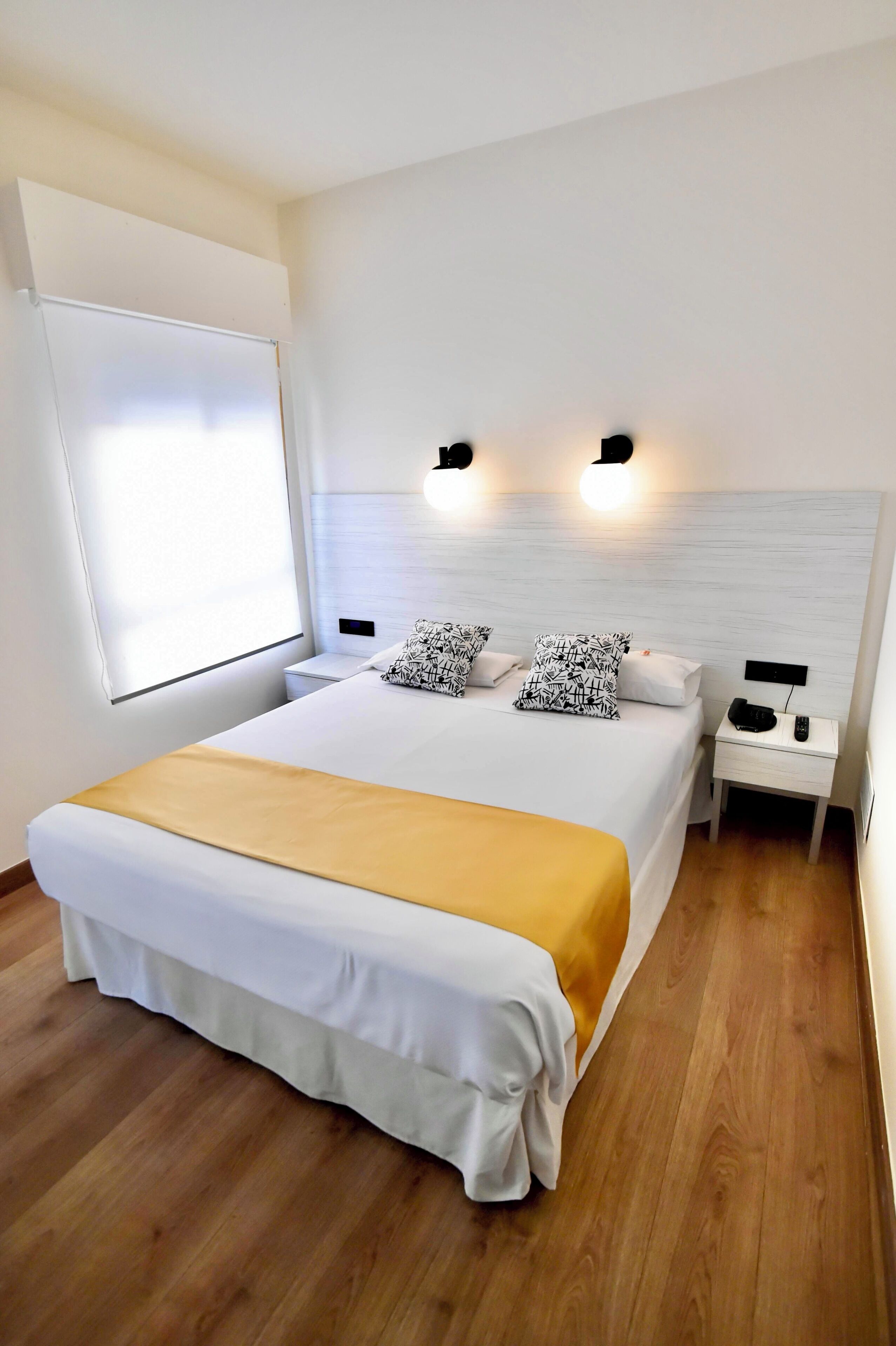 A hotel room with a large bed, bedside tables, a window, and wall-mounted lights.