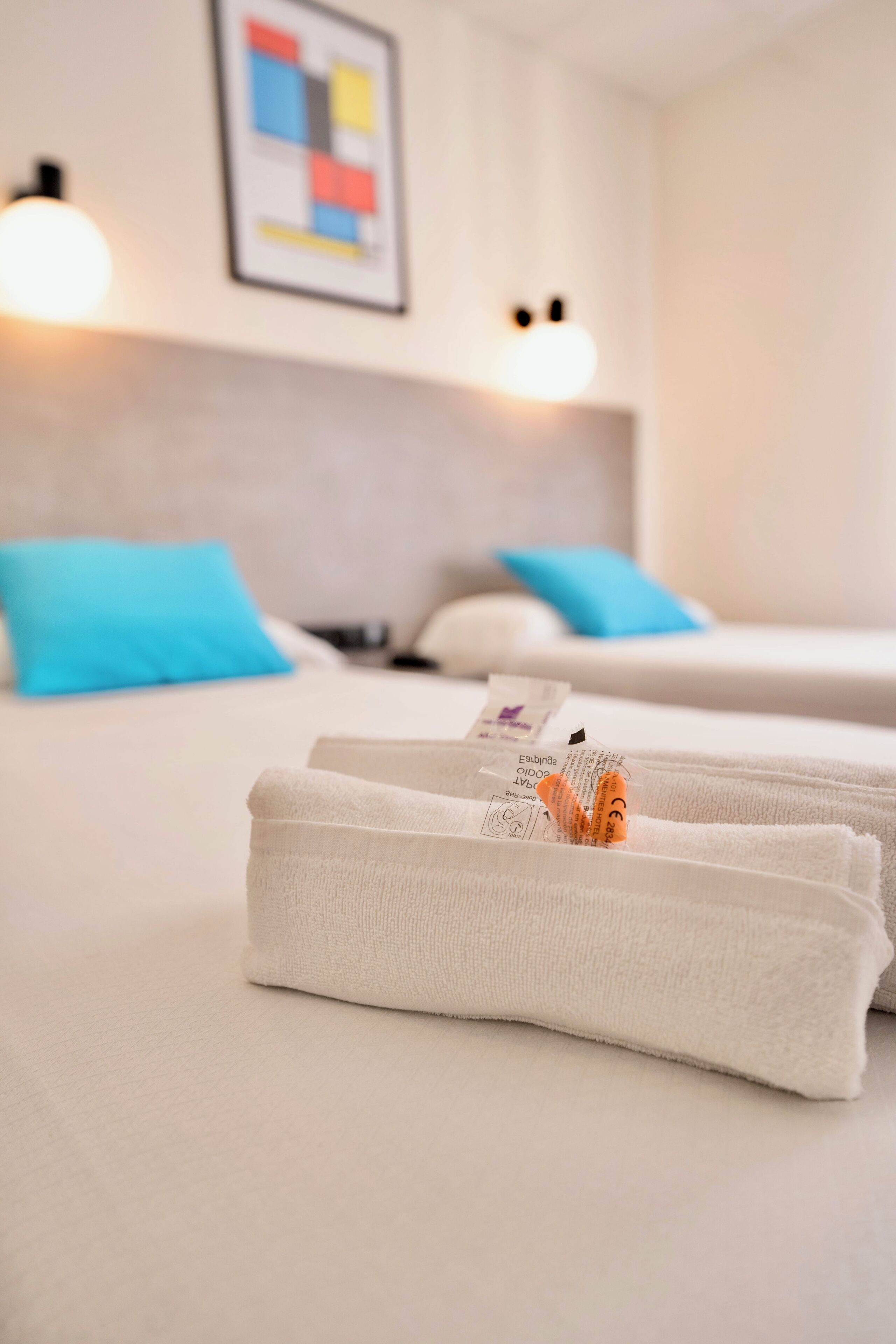 Habitación de hotel con dos camas, cada una con una almohada azul, y una toalla blanca doblada cuidadosamente sobre la cama.