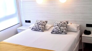 Quarto de hotel com cama, duas mesas de cabeceira, um criado-mudo e luminária fixada na parede.