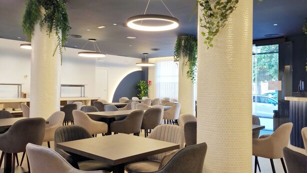 Uma sala de jantar moderna com mesas e cadeiras redondas, luminárias decorativas no teto e plantas verdes em colunas.