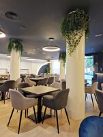 A modern dining area with round tables and chairs, decorative ceiling lights, and green plants on columns.