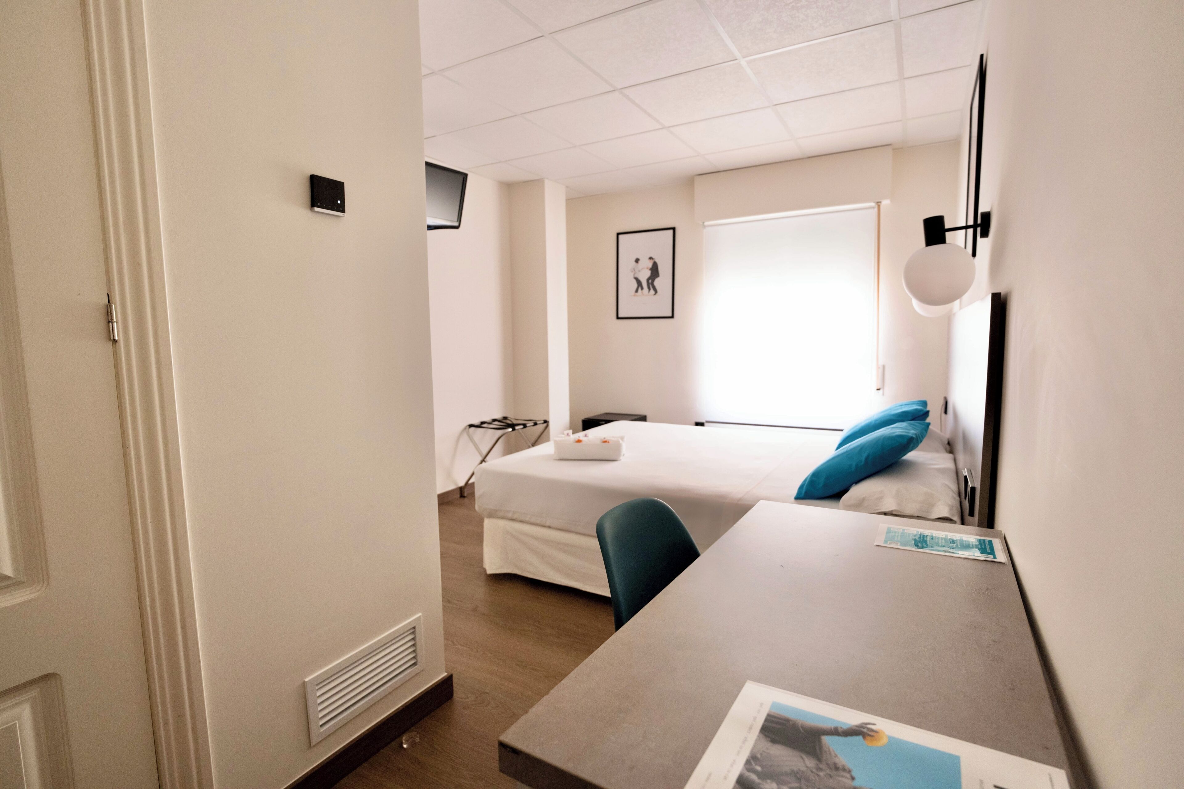 A hotel room with a bed, a desk, a chair, a window, and a wall-mounted light fixture.