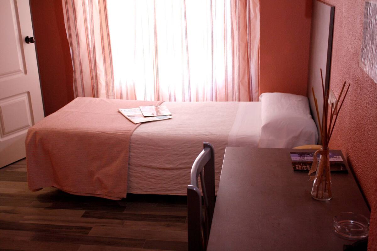 A hotel room with a bed, a chair, a table, and a vase with reed sticks.