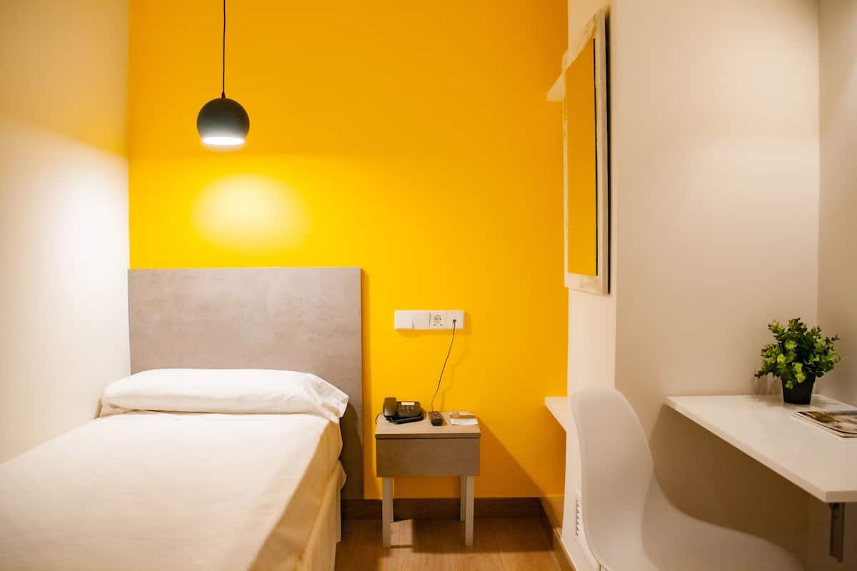 A hotel room with a bed, bedside table, desk, chair, and a potted plant.