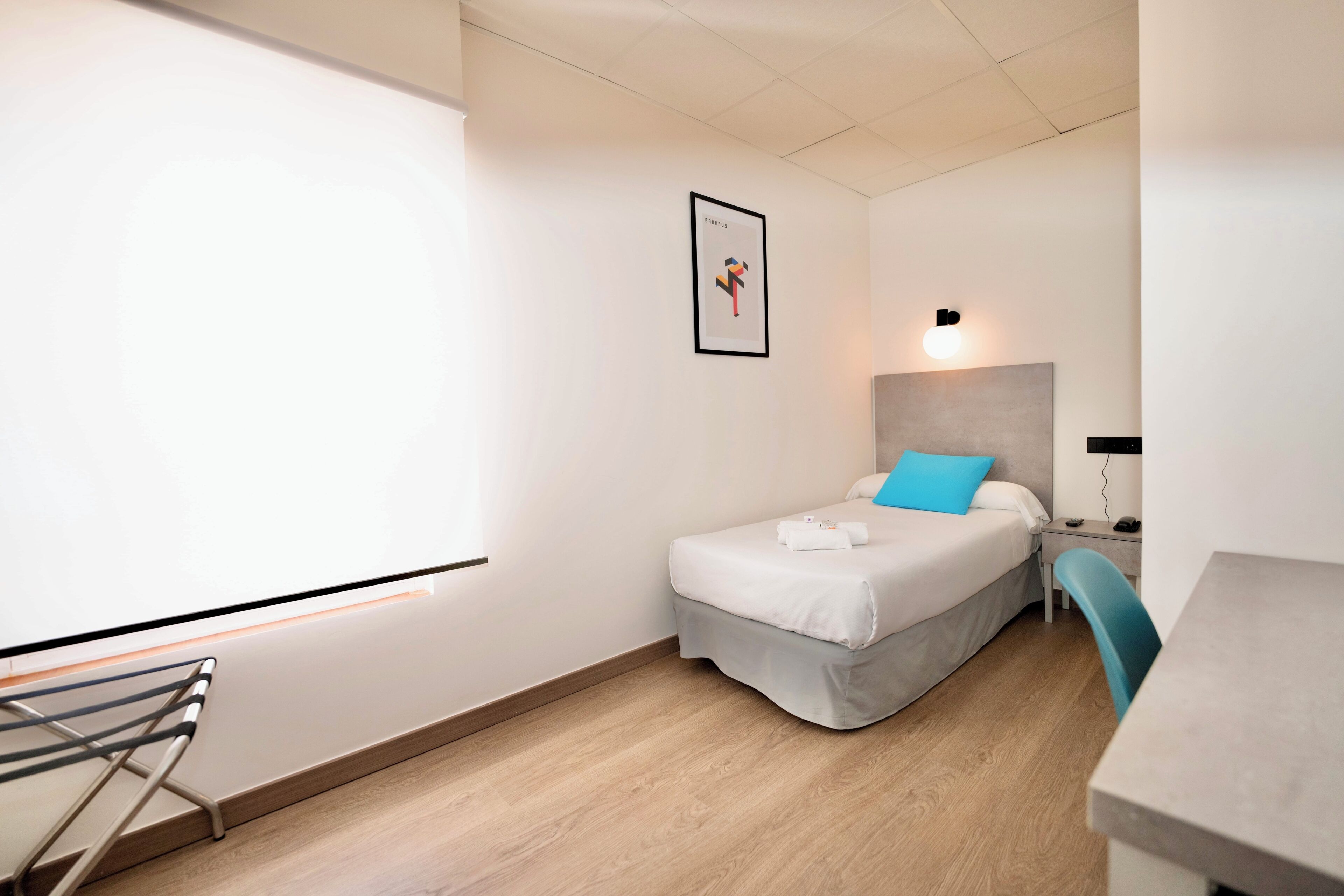 A modern hotel room with a bed, a desk, a chair, a wall-mounted projector, and a framed picture.