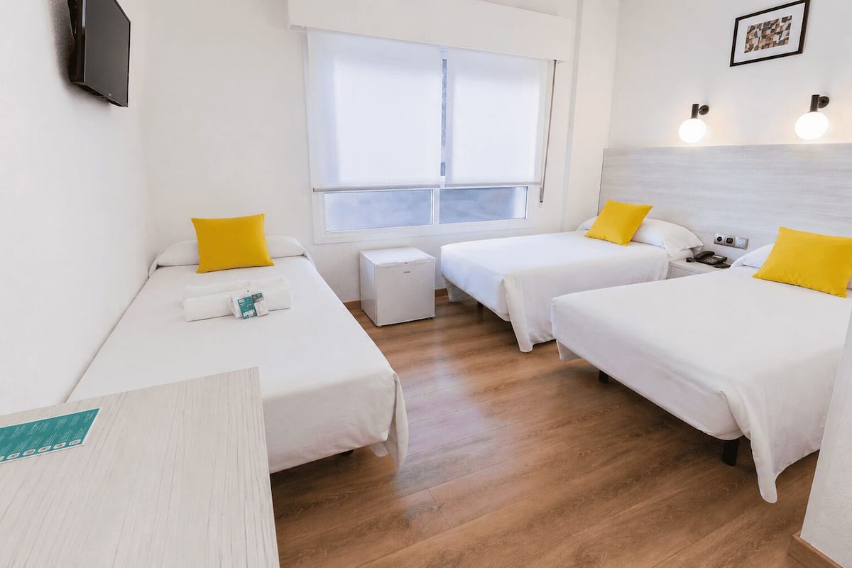 A hotel room with two single beds, a small table, a bedside table with a phone, a window with blinds, and a wall-mounted TV.