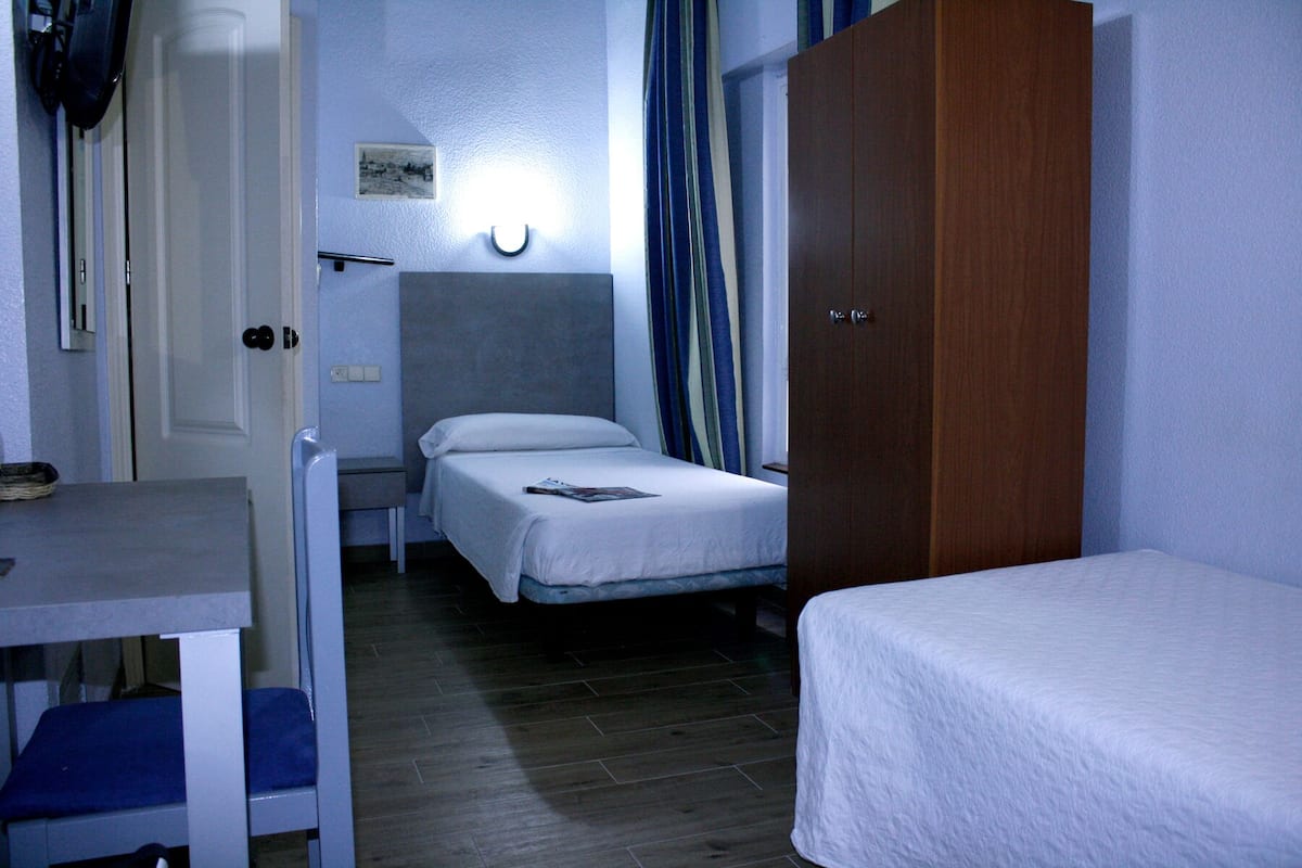 A compact hotel room with two single beds, a wooden wardrobe, a small desk with a chair, and a bedside table.