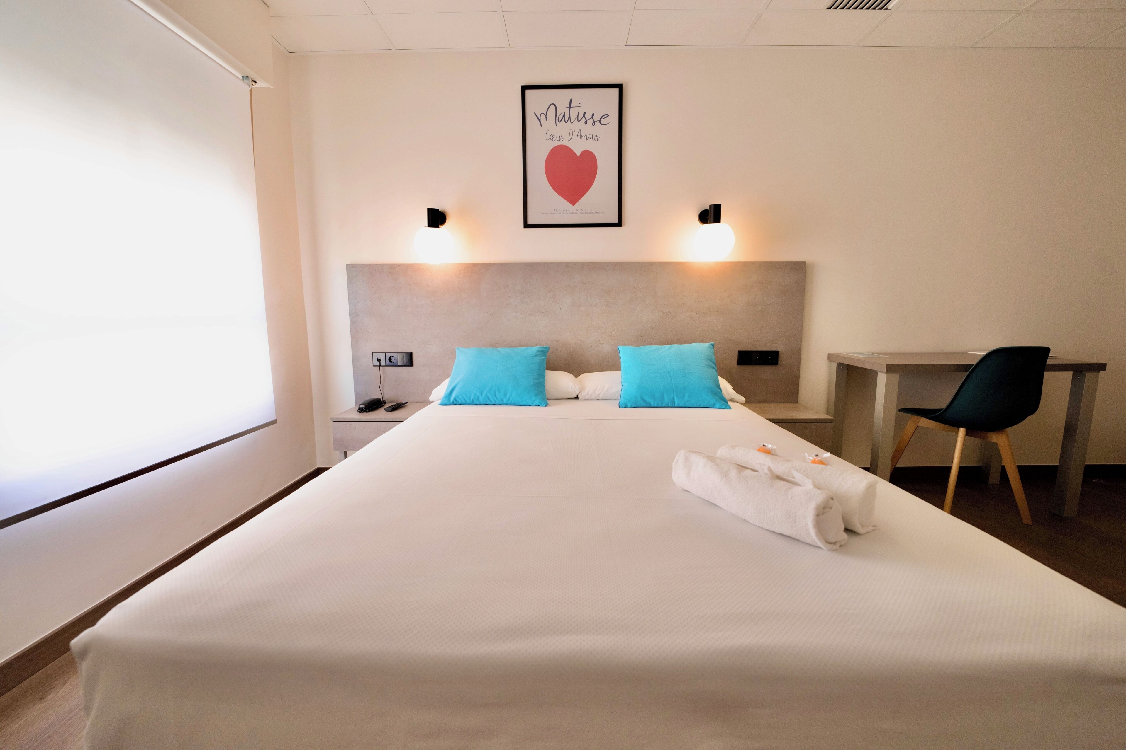 Quarto de hotel com cama, duas almofadas, escrivaninha, cadeira, mesa de cabeceira e uma moldura na parede.