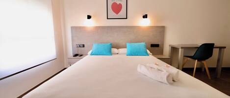 Una habitación de hotel con una cama, dos almohadas, un escritorio, una silla, una mesita de noche y un cuadro en la pared.