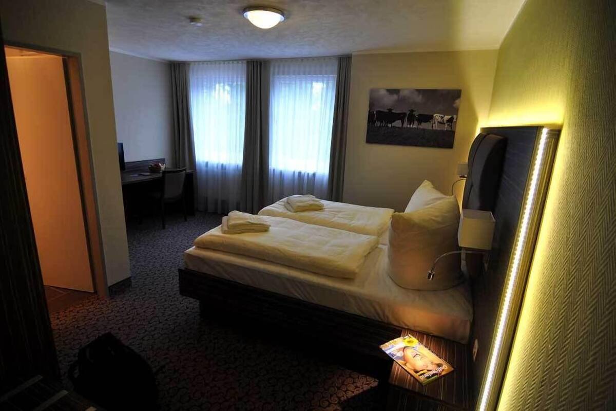A hotel room with a bed, a desk, and a chair. There is a window with curtains, a wall-mounted lamp, and a picture on the wall.