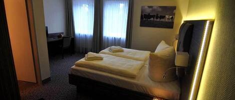 A hotel room with a bed, a desk, and a chair. There is a window with curtains, a wall-mounted lamp, and a picture on the wall.