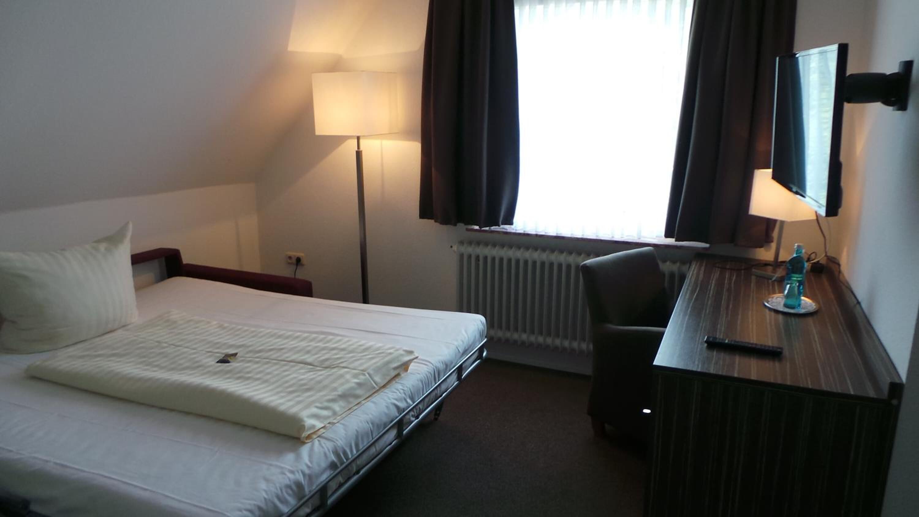Hotelkamer met een bed, een bureau, een stoel, een lamp, een radiator en een raam met gordijnen.