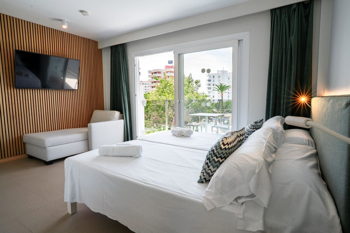 A modern hotel room with a large bed, a TV mounted on the wall, a balcony with a view of the city, and a seating area with a sofa and a table.