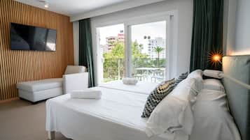 A modern hotel room with a large bed, a TV mounted on the wall, a balcony with a view of the city, and a seating area with a sofa and a table.