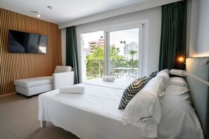 A modern hotel room with a large bed, a TV mounted on the wall, a balcony with a view of the city, and a seating area with a sofa and a table. - INN Mallorca Aparthotel (Calvia)