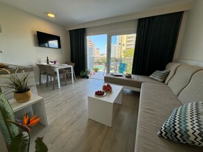 A modern living room with a sofa, coffee table, dining table, and chairs. There is a TV mounted on the wall and a balcony with a view of buildings. - INN Mallorca Aparthotel (Calvia)