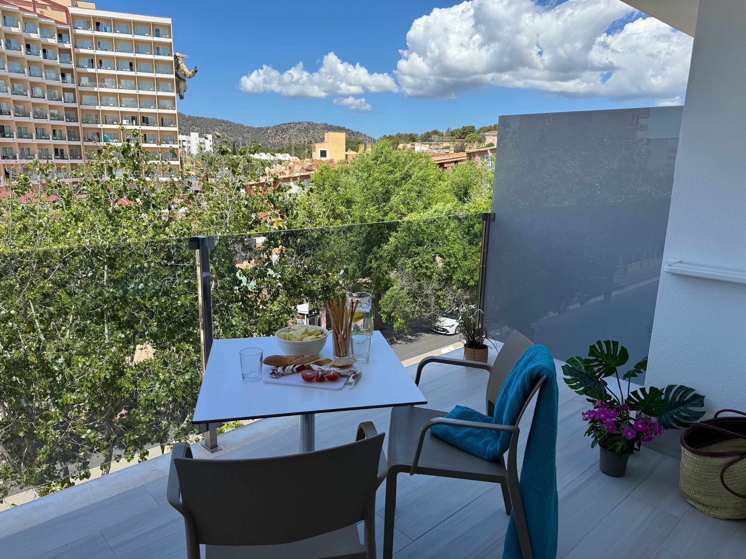 Un balcón con una mesa dispuesta para dos, una silla, una planta en maceta y vistas a un paisaje urbano con edificios y árboles.