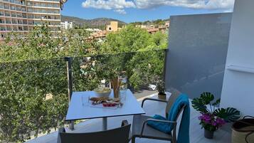 Un balcón con una mesa dispuesta para dos, una silla, una planta en maceta y vistas a un paisaje urbano con edificios y árboles.