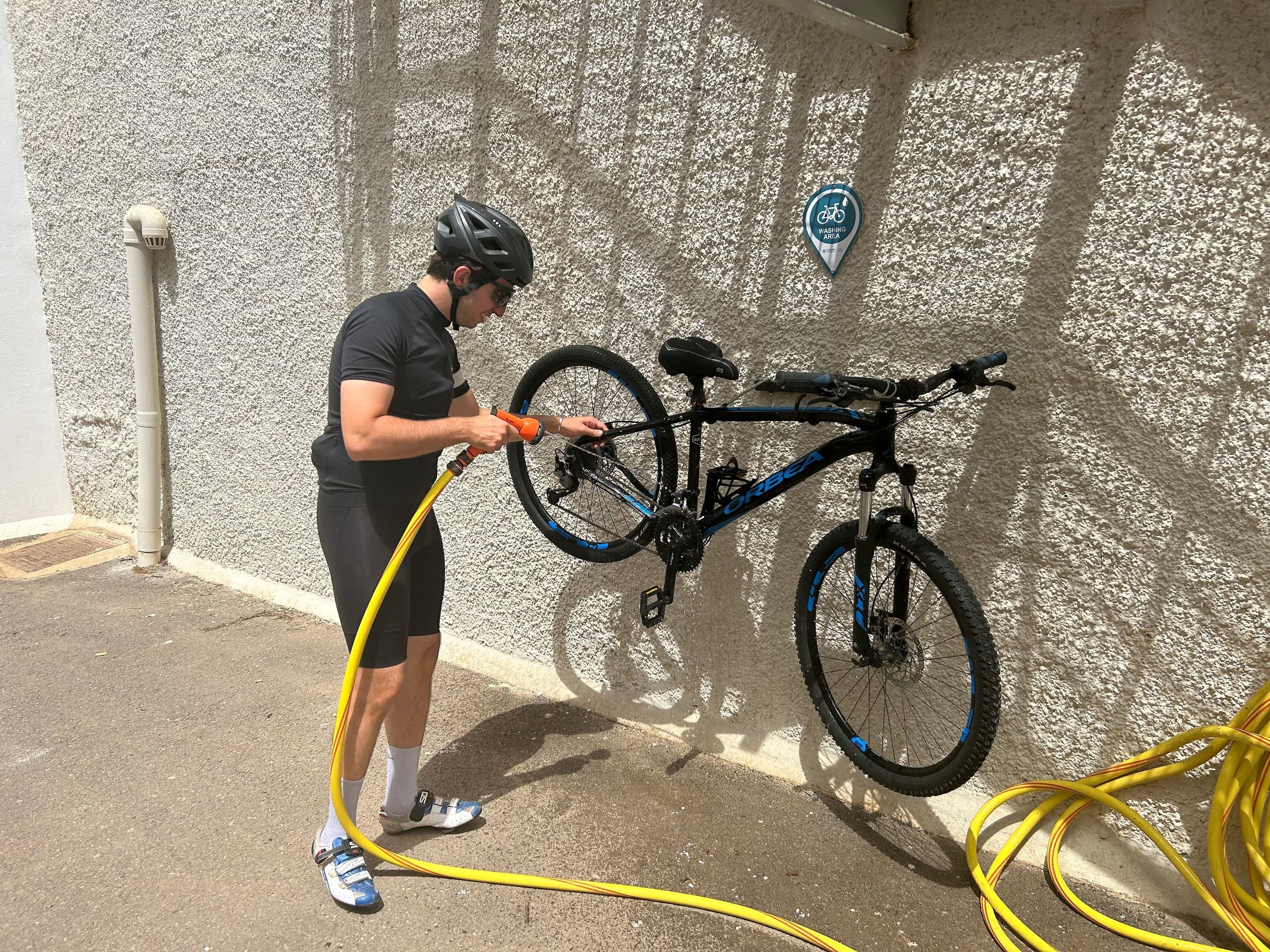 Una persona limpia una bicicleta con una manguera amarilla frente a una pared con textura.