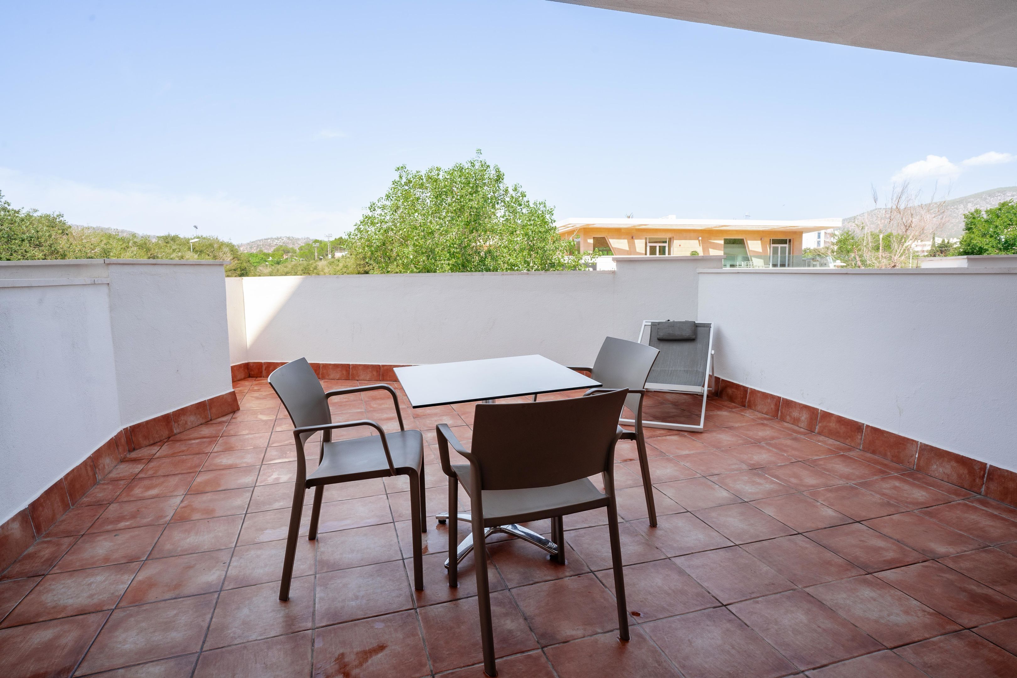 An outdoor patio with a table and chairs, a tiled floor, and a view of a building and trees in the background.