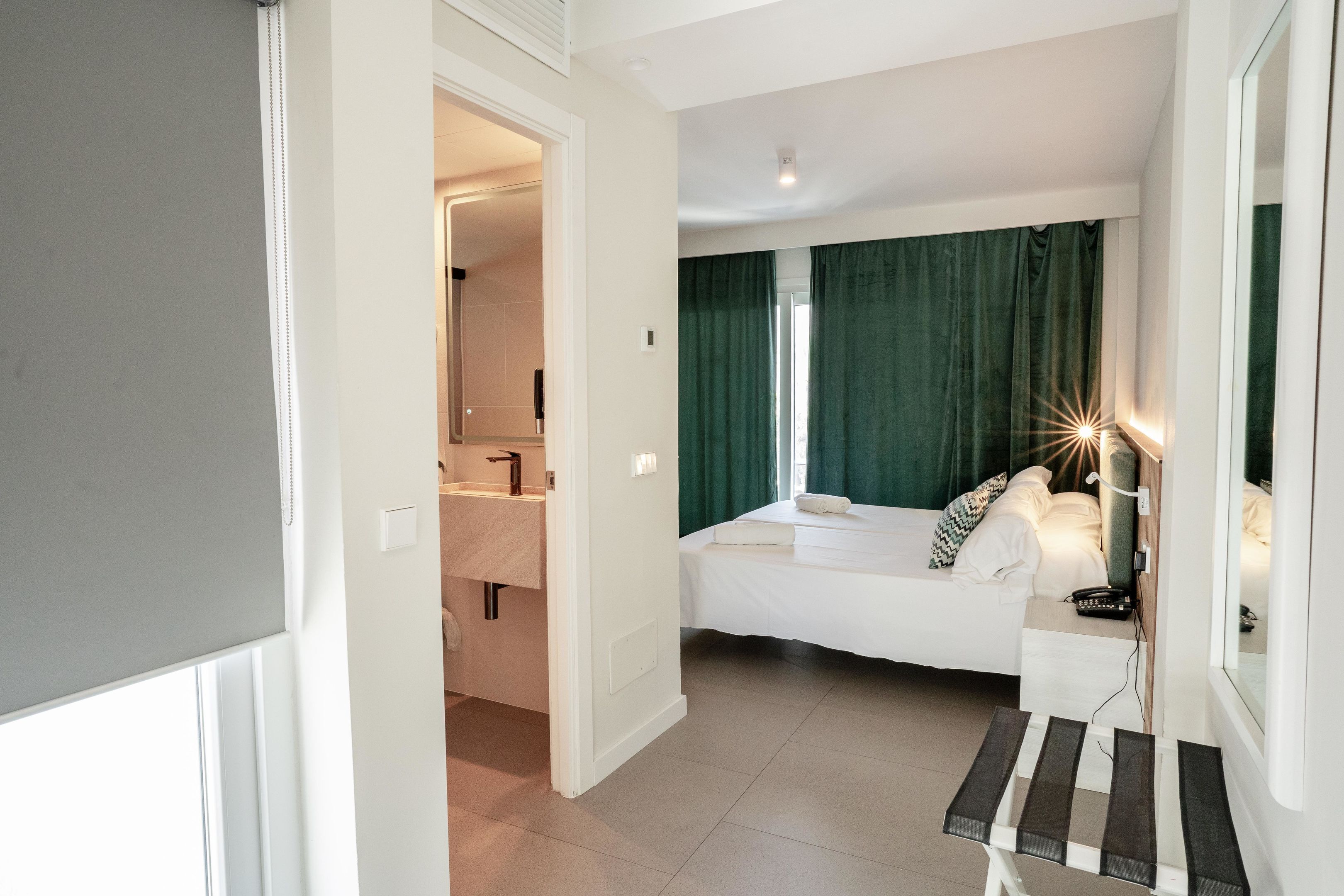 Un dormitorio con cama, mesita de noche y espejo. Se ve un baño con lavamanos y espejo.