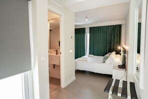 A bedroom with a bed, bedside table, and a mirror. There is a bathroom visible with a sink and mirror.