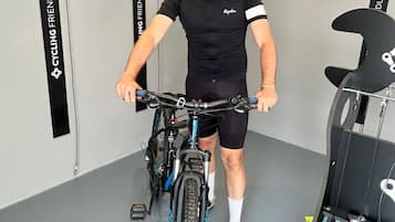 Una persona en una bicicleta estática en una habitación con piso y paredes grises, una pared a rayas negras y blancas, y una pared negra con una franja blanca y texto.