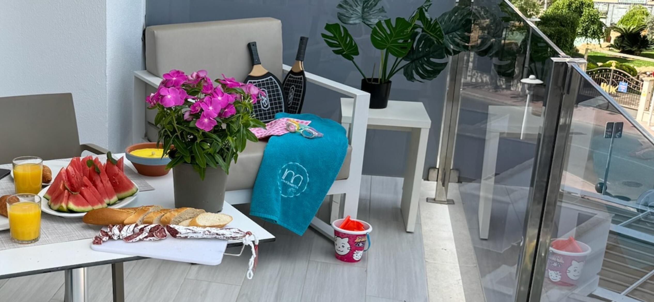 Un balcon avec une table préparée pour le petit-déjeuner, comprenant de la pastèque, du pain et du jus de fruits.
