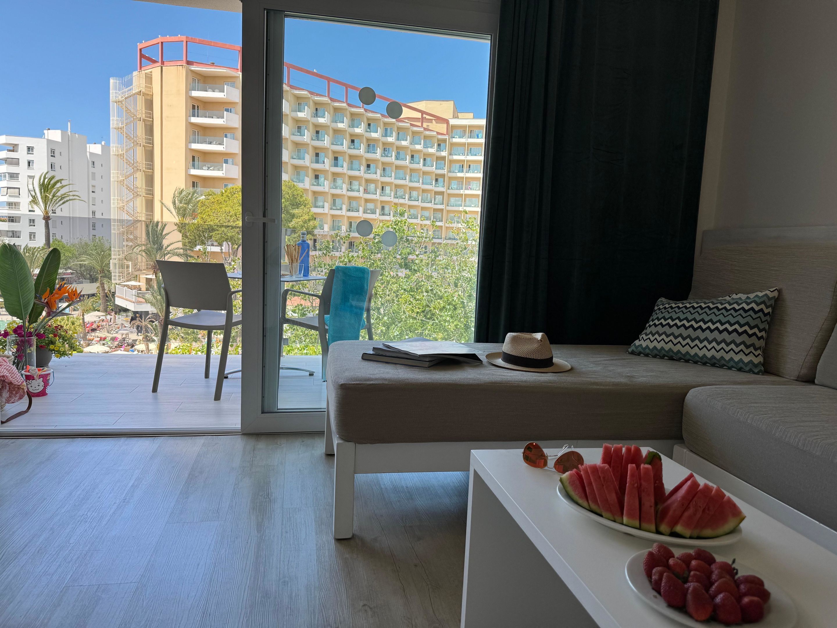 Habitación de hotel moderna con balcón, sofá, mesa de centro con fruta y vista a la ciudad.