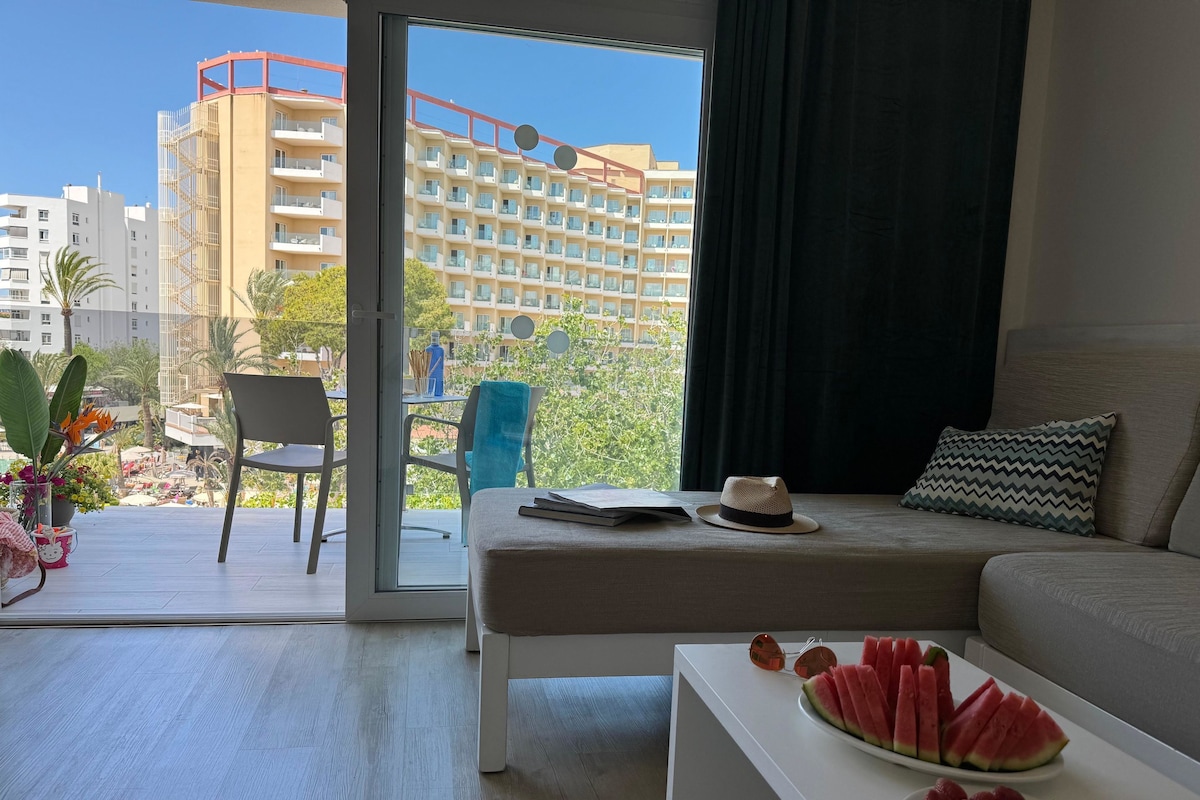 A modern hotel room with a balcony, a sofa, a coffee table with fruit, and a view of the cityscape.
