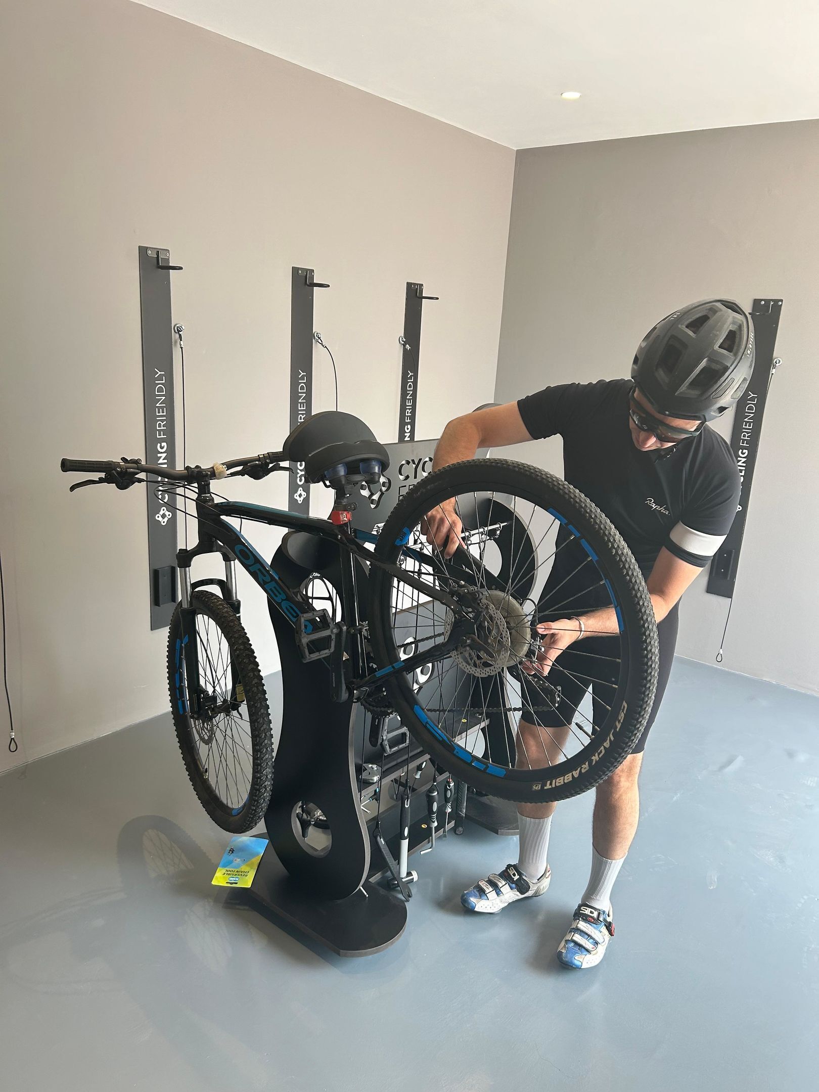 Iemand past een fiets op een trainingsstandaard in een ruimte met een grijze vloer en witte muren.