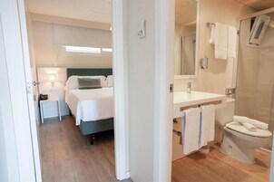 Moderni hotellihuone, jossa on sänky, yöpöytä, lamppu sekä kylpyhuone, jossa on pesuallas, peili ja wc.
