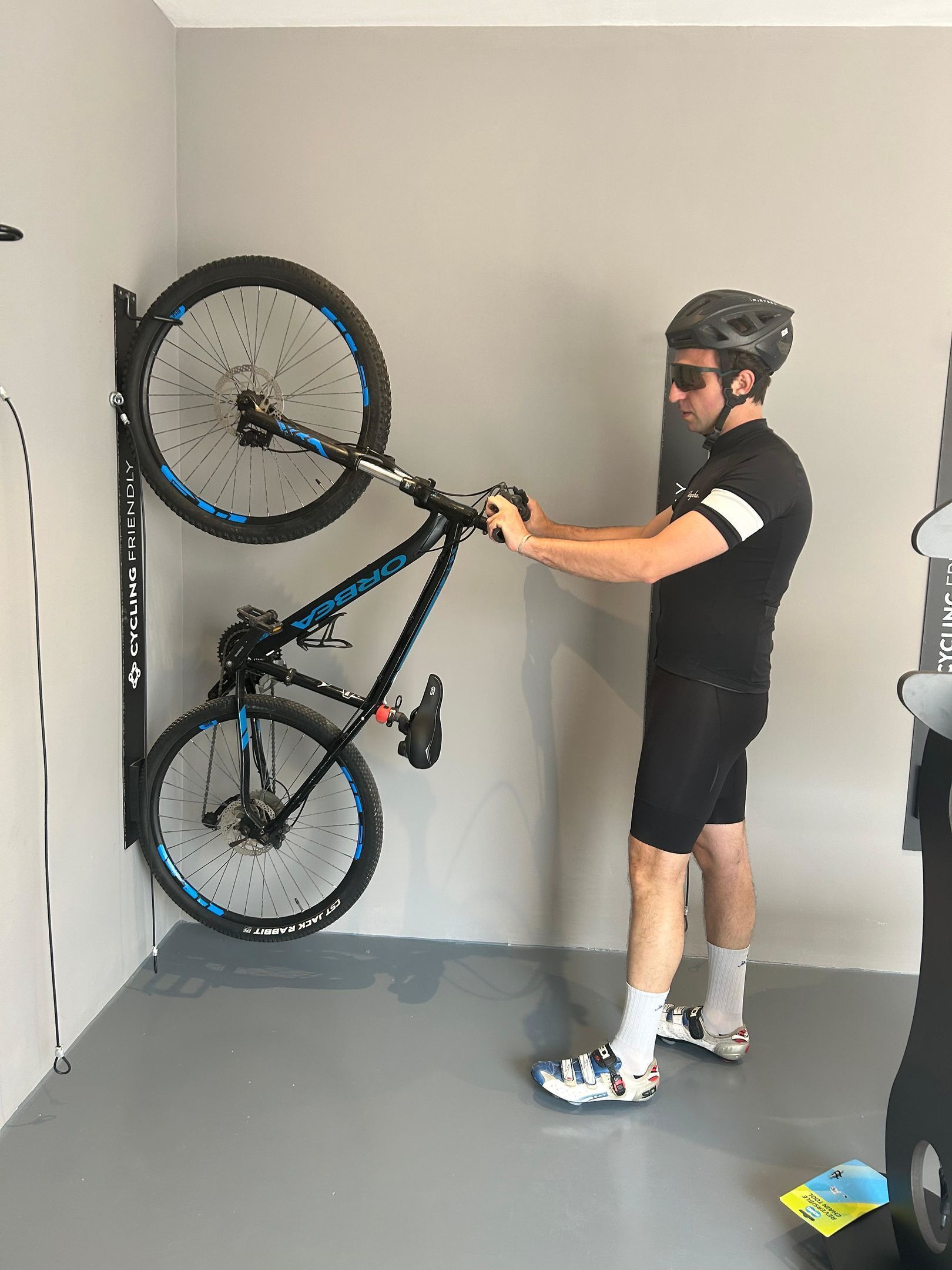 Une personne est en train de régler un vélo dans une pièce dont le sol et les murs sont gris, où l'on aperçoit un support à vélos fixé au mur et un tapis de course.