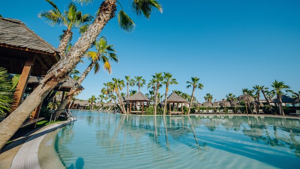 A resort pool with palm trees, thatched-roof huts, and lounge chairs. -  Alannia Costa Blanca Resort (Crevillente)