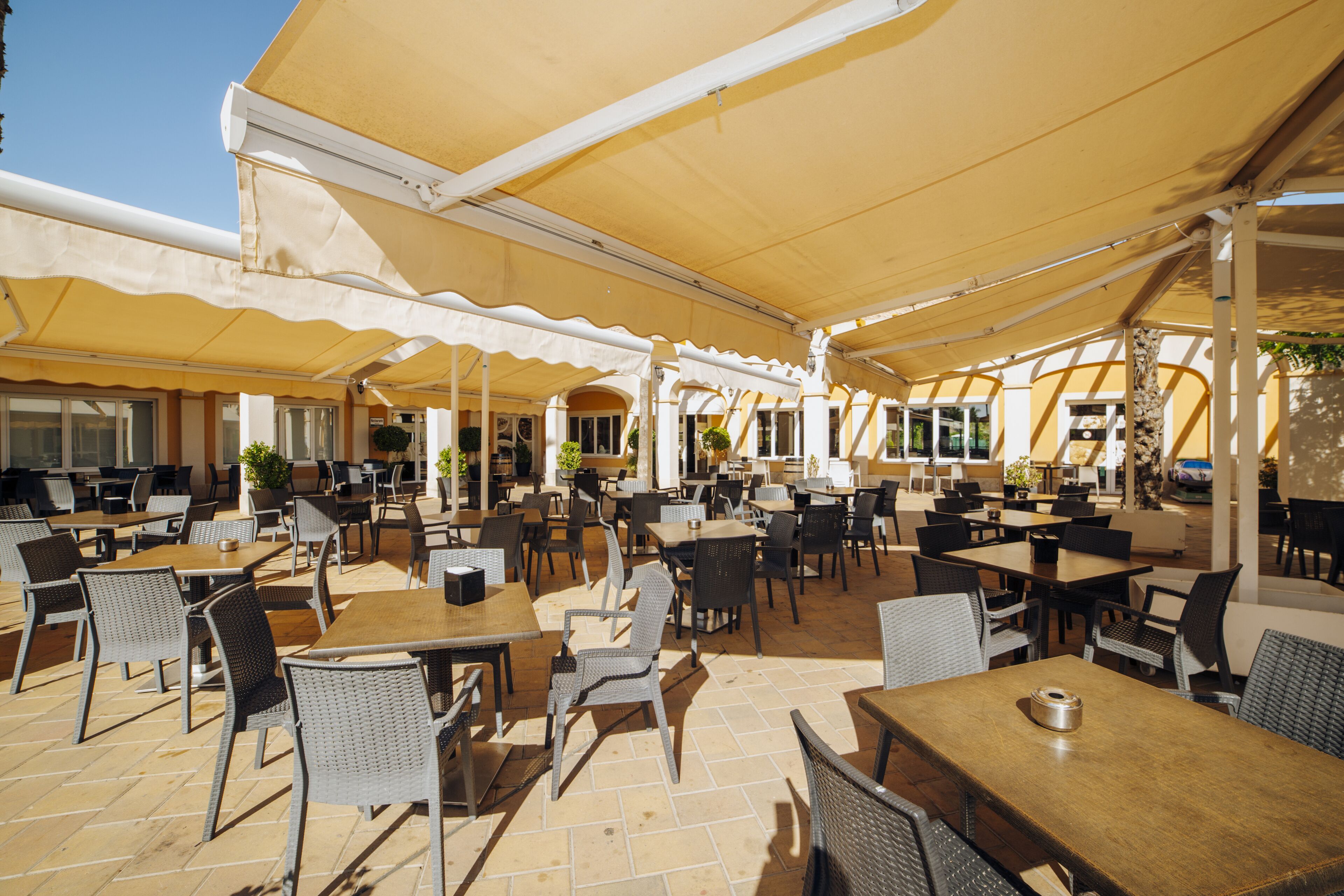 An outdoor café with numerous tables and chairs, covered by large umbrellas, and surrounded by potted plants.