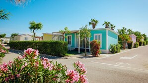 A row of colorful mobile homes with a clear sky, a road with a directional arrow, and flowering plants in the foreground. -  Alannia Costa Blanca Resort (Crevillente)