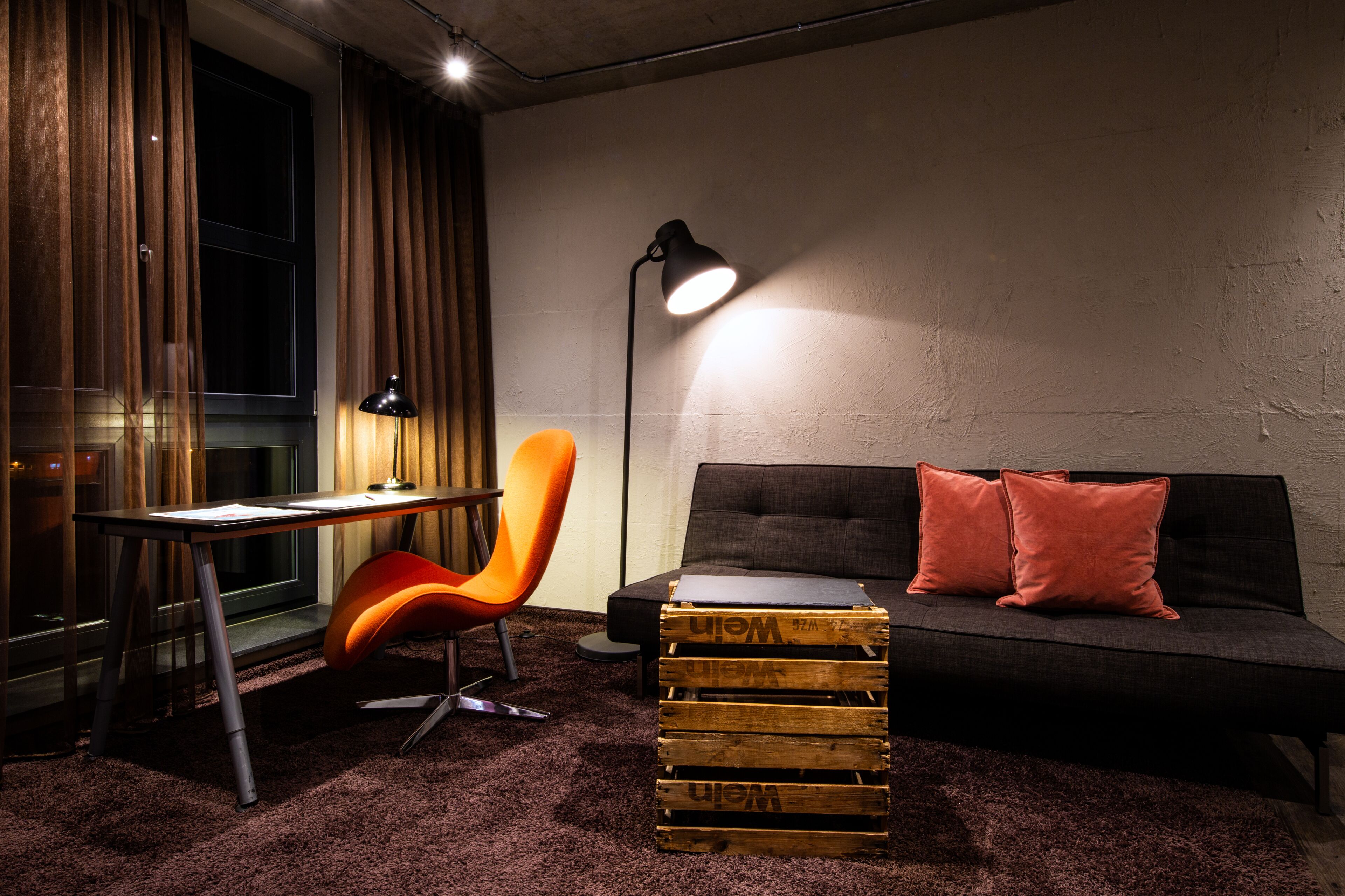 A room with a desk, an orange chair, a black sofa with red cushions, and a lamp.