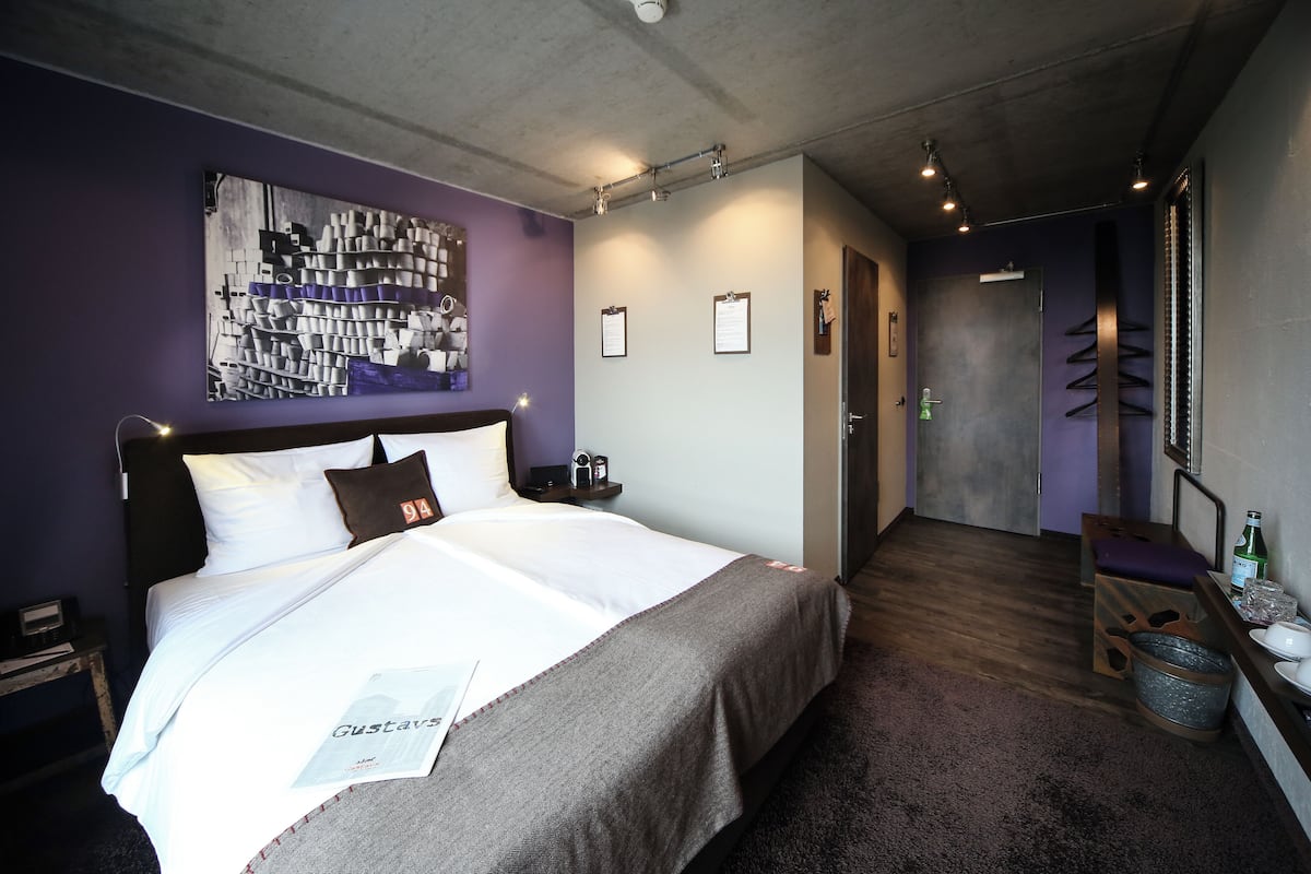 Quarto de hotel moderno com uma cama grande, cabeceira, obra de arte abstrata na parede, mesinha de cabeceira, criado-mudo, um banco de madeira, um cesto de lixo e uma porta que dá para outro quarto.