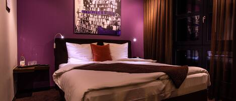 A neatly made bed with white linens and a brown throw, a nightstand with a bottle and glasses, and a large abstract painting on the purple wall.