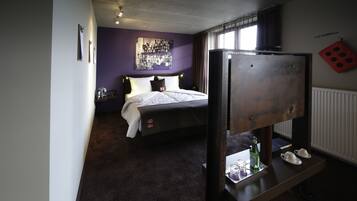 A hotel room with a bed, a small table with a bottle and glasses, a wall-mounted TV, and a window with curtains.