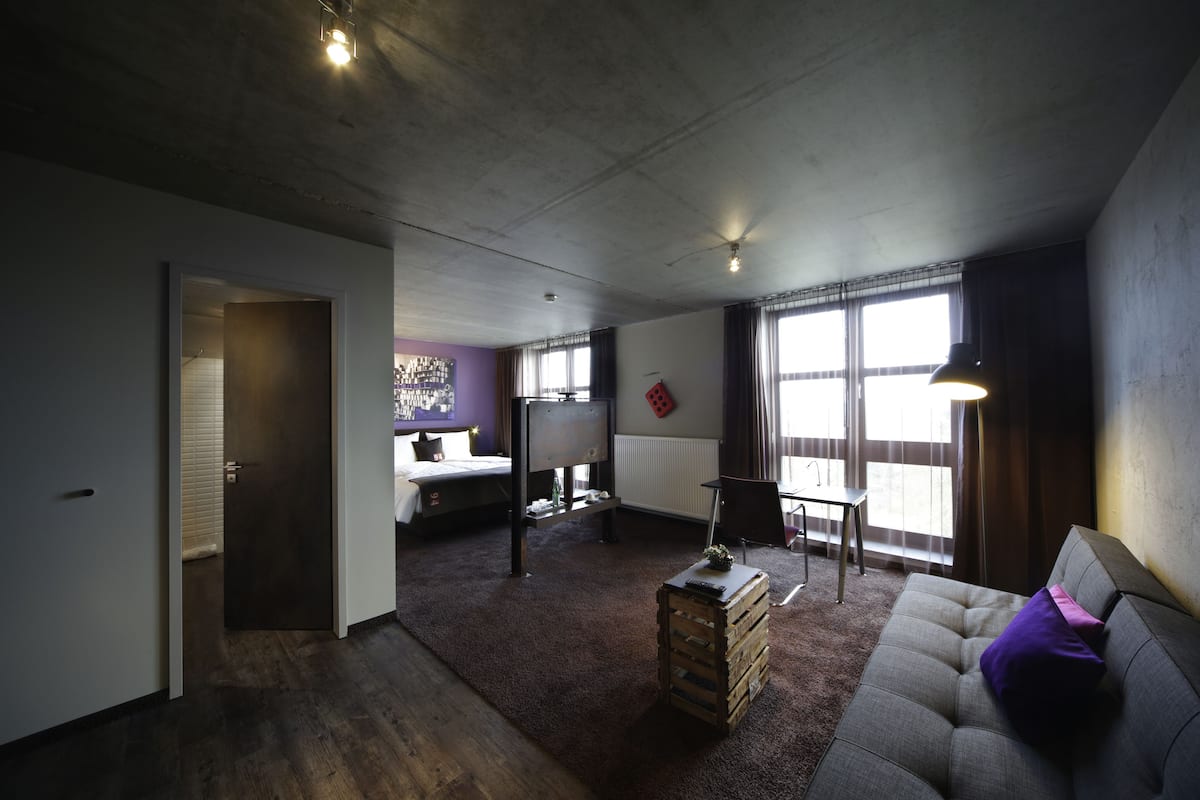 Uma sala de estar moderna com um sofá, uma mesa de centro de madeira e uma televisão. Há uma porta que dá para outro cômodo, e uma janela com cortinas.